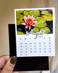 A person's hand holding a small calendar with a photo of a pink flower on a pond.