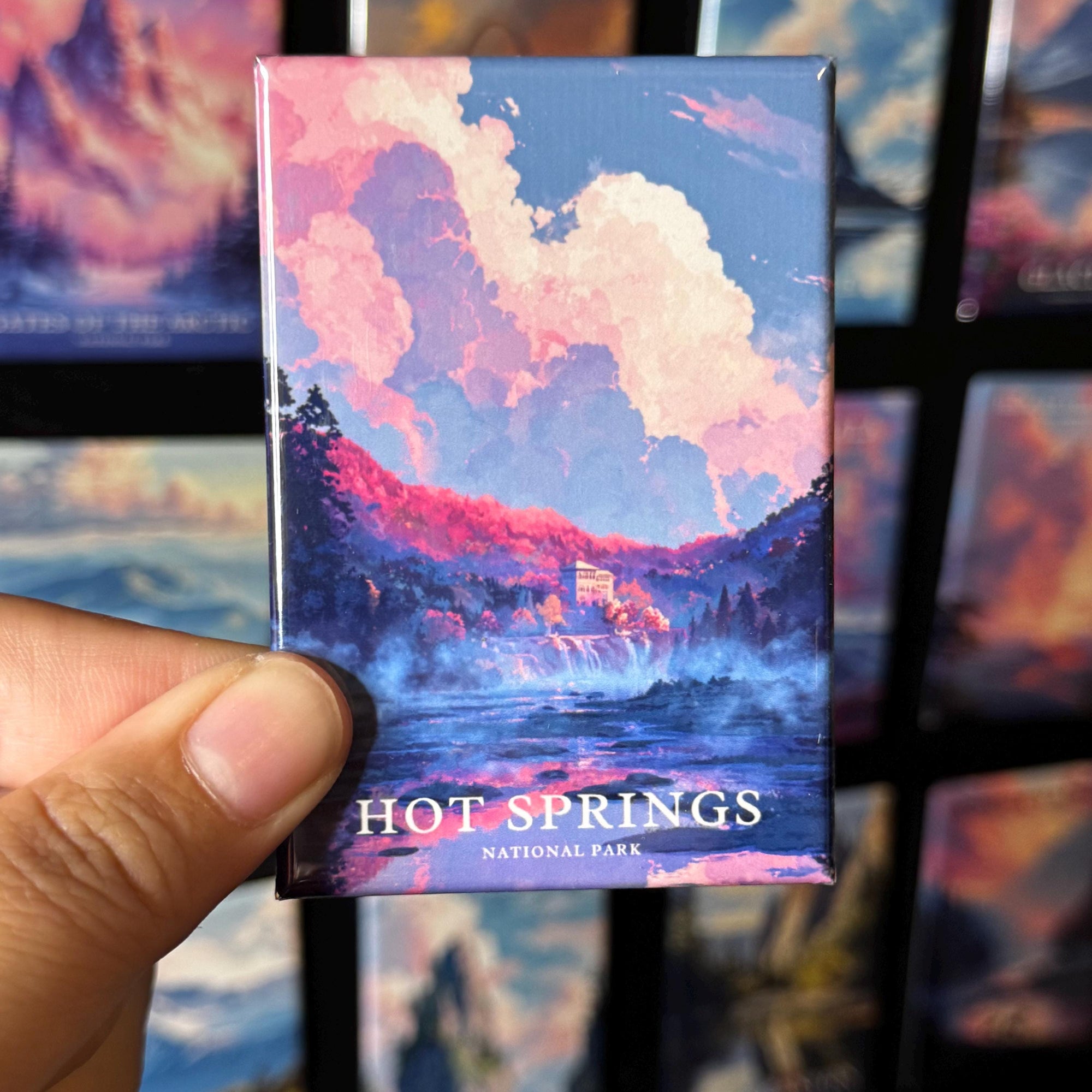 A hand holding a magnet with a colorful illustration of Hot Springs National Park, featuring a scenic landscape with a waterfall and clouds.