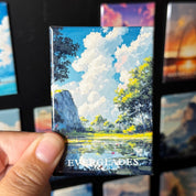 A hand holding a magnet with a painting of a landscape featuring a lake, trees, and a cloudy sky.