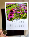 A hand holding a calendar card with a photo of a field of purple flowers.