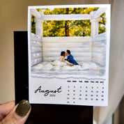 A person is holding a calendar for August 2026, featuring a photo of a couple embracing in a white wooden structure surrounded by trees.
