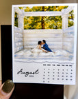 A person is holding a calendar for August 2026, featuring a photo of a couple embracing in a white wooden structure surrounded by trees.