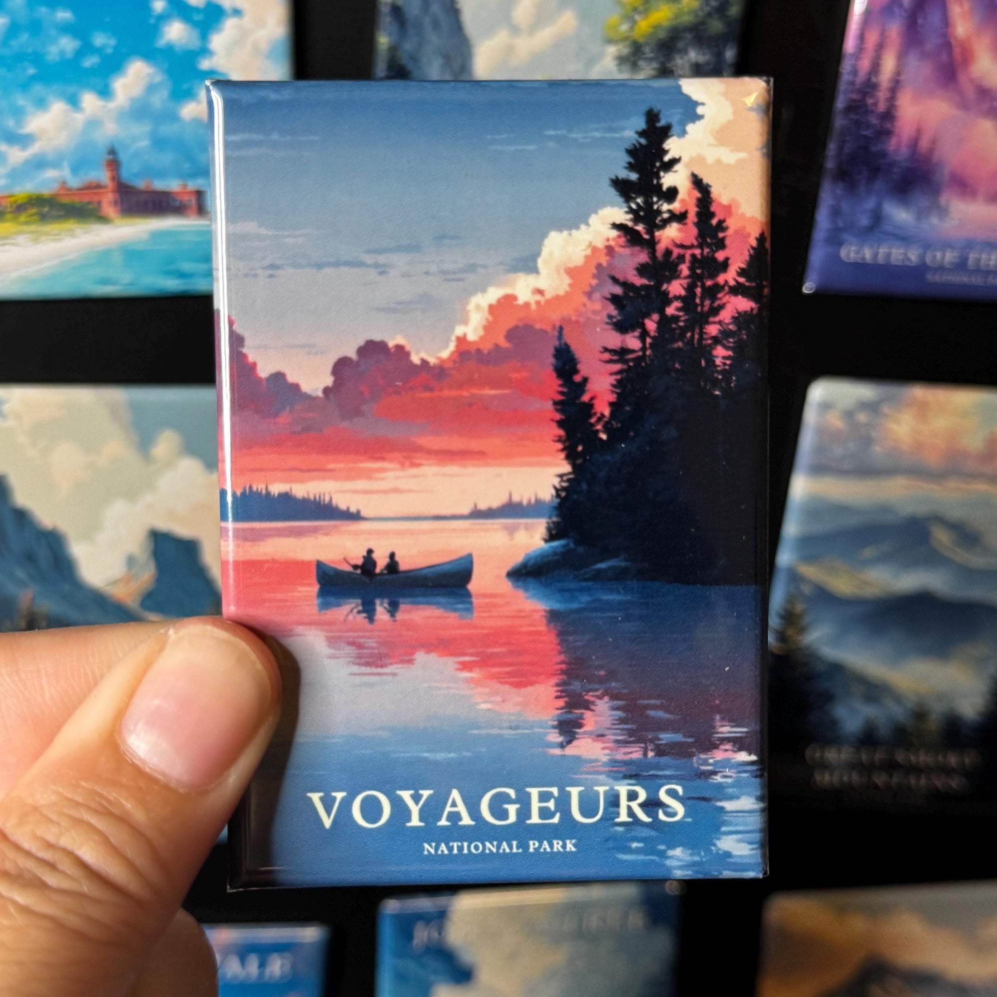 A hand is holding a magnet with a colorful illustration of a boat on a lake, surrounded by trees and a sunset sky.