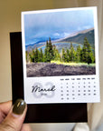 A hand holding a small calendar with a photo of a forest and mountains on the cover, and a calendar for the month of March 2016.