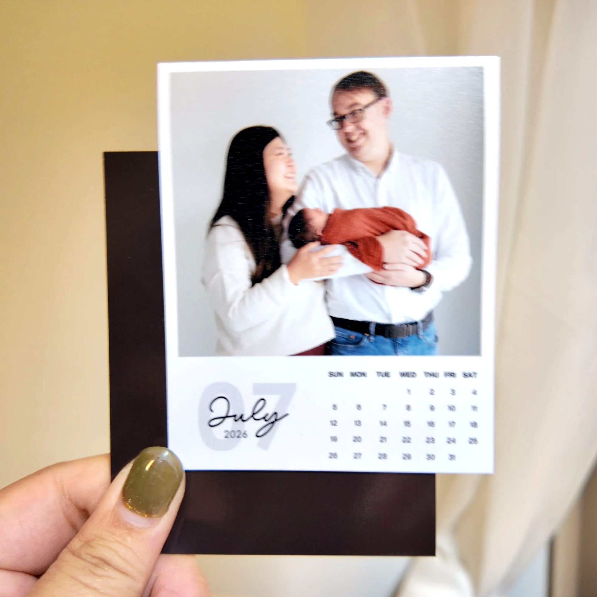 A person is holding a small calendar featuring a photo of a couple with a baby, with the date "July 2026" visible.