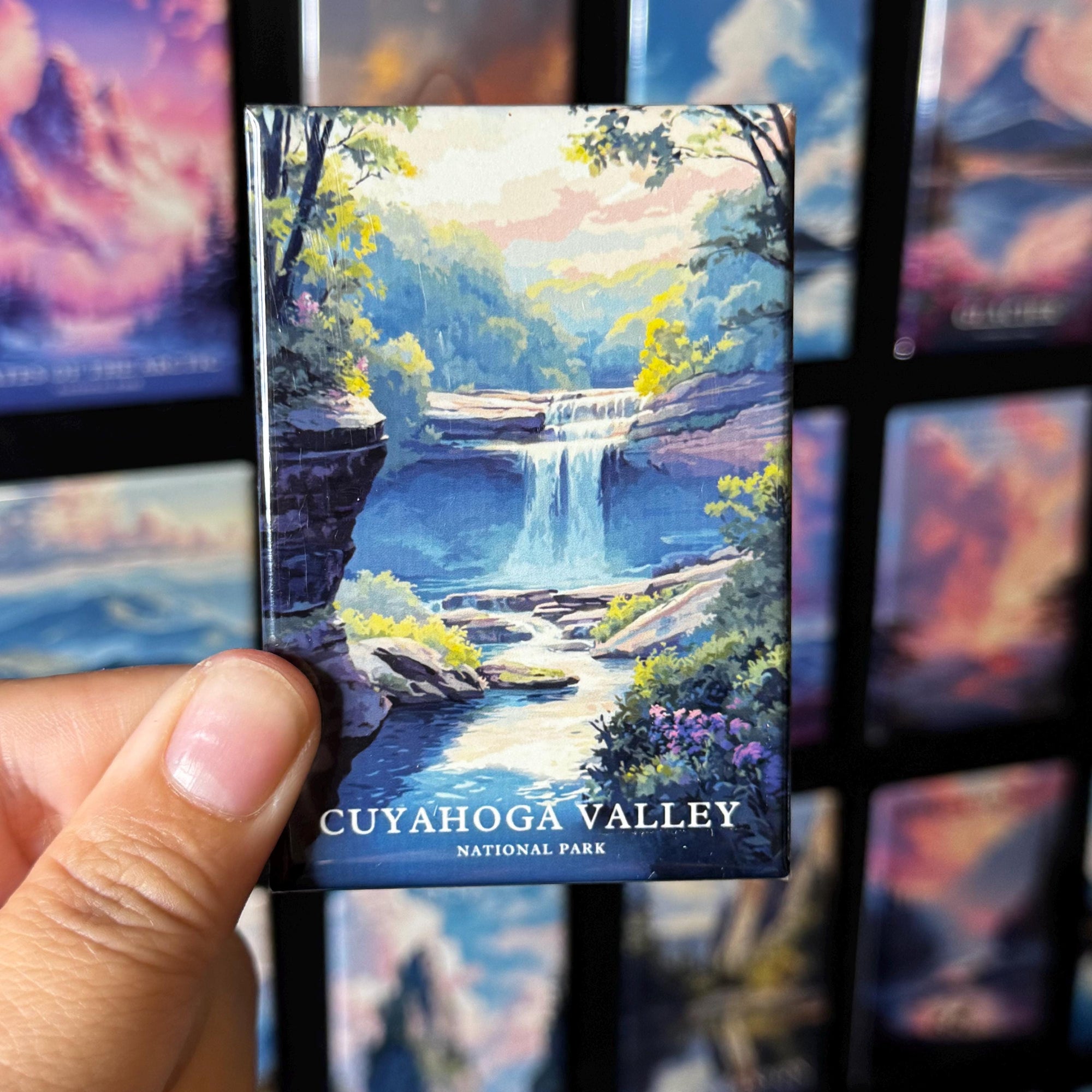 A hand holding a magnet with a painting of a waterfall and forest in the Cuyahoga Valley National Park.