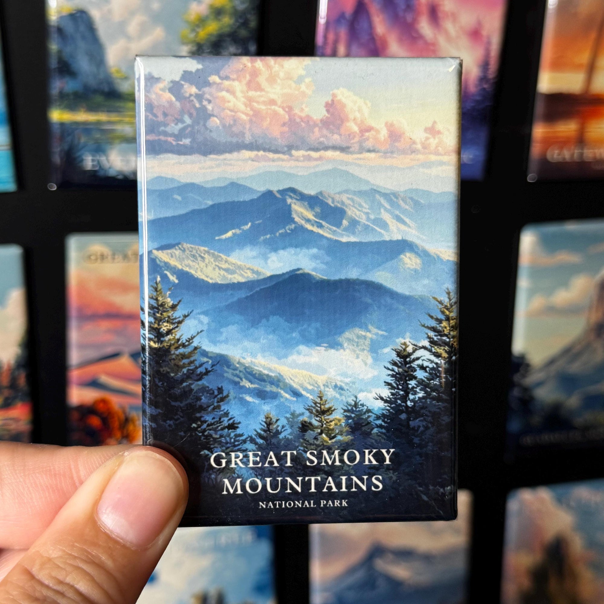 A hand holding a magnet with a scenic mountain landscape featuring clouds and pine trees.