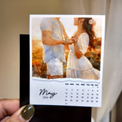 A hand holding a small calendar with a photo of a couple on the cover, and a calendar for the month of May.