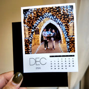 A person is holding a small photo album with a photo of a family standing in front of a beautifully decorated archway, surrounded by twinkling lights.