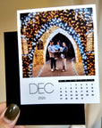 A person is holding a small photo album with a photo of a family standing in front of a beautifully decorated archway, surrounded by twinkling lights.