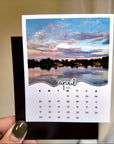 A hand holding a calendar card with a photo of a lake and a sunset.