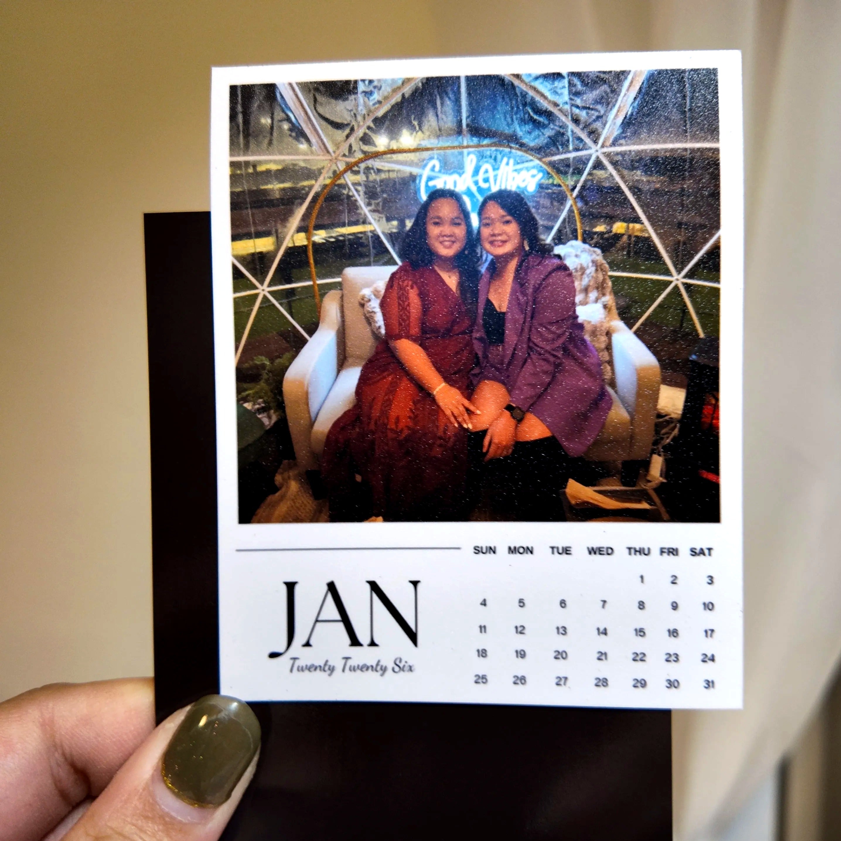 A person is holding a photo of two women sitting on a couch, with a calendar for the month of January visible.