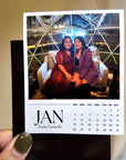A person is holding a photo of two women sitting on a couch, with a calendar for the month of January visible.