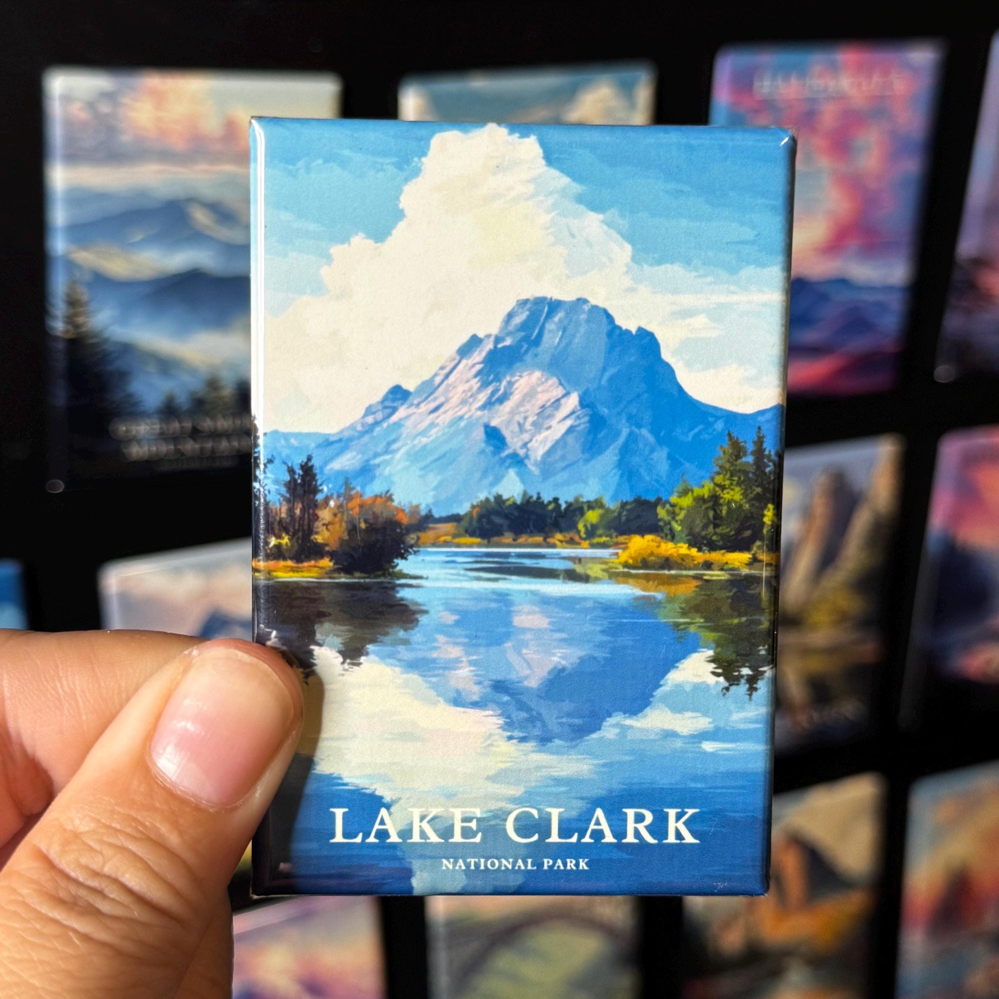A hand is holding a magnet with a painting of Lake Clark and its surroundings, including a mountain and a lake, against a background of other landscape paintings.
