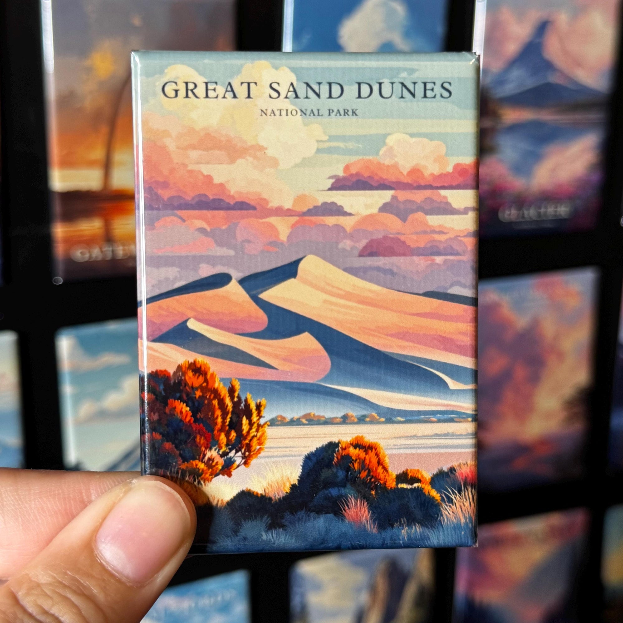 A hand holding a magnet with an illustration of sand dunes and mountains, with the text "GREAT SAND DUNES NATIONAL PARK" printed on it.