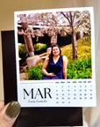 A person holding a photo of a young woman posing in front of a cherry blossom tree, with a calendar for the month of March visible.