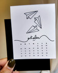 A person is holding a calendar card with a simple design of paper airplanes on it.