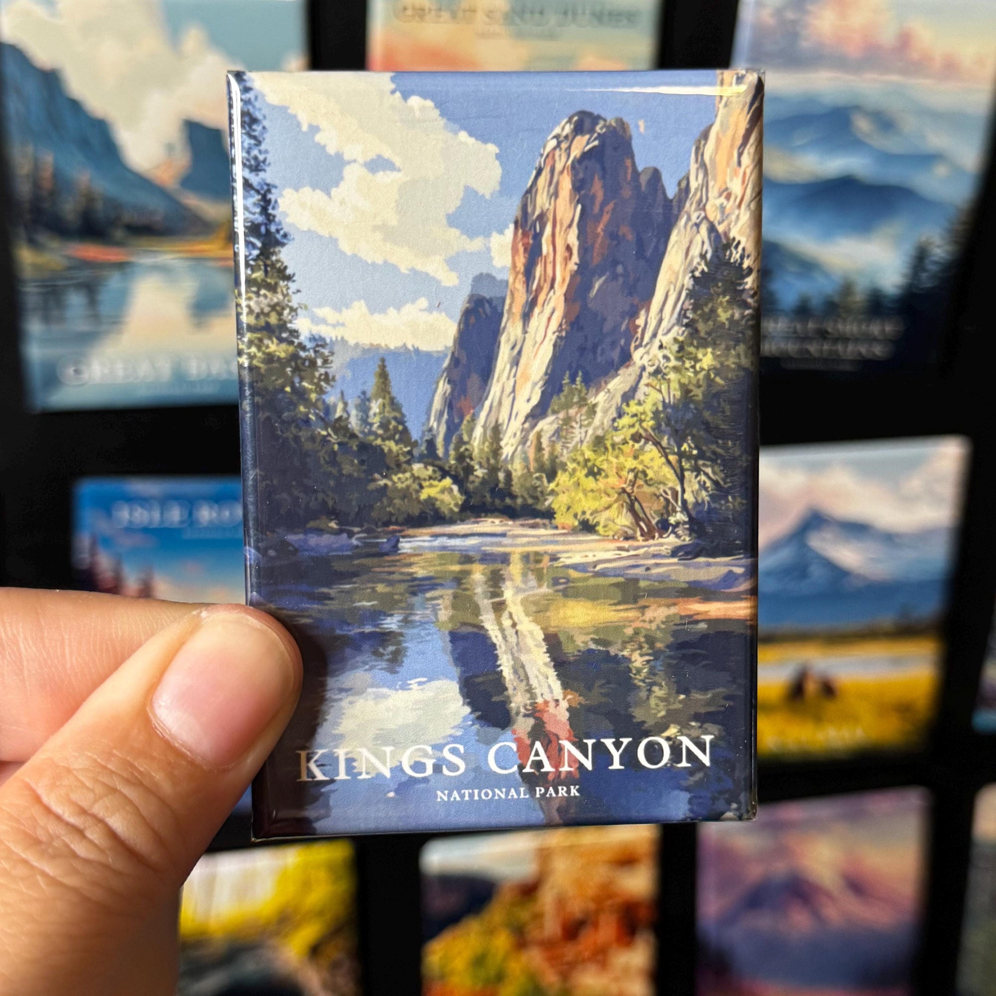 A hand is holding a magnet with a scenic image of the Kings Canyon National Park, featuring a mountain landscape with a lake and trees.