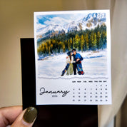 A person is holding a small calendar with a photo of a couple embracing in front of a snowy mountain landscape.