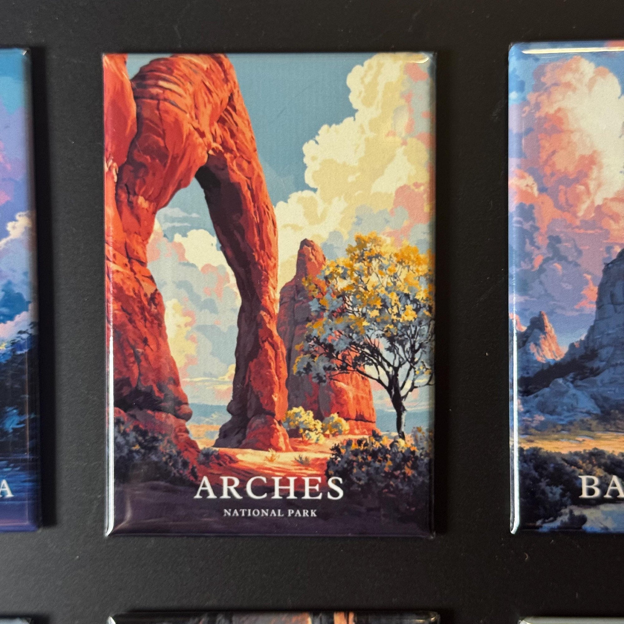 a collection of landscape magnets, including a prominent magnet with an illustration of a large arch formation surrounded by trees and a body of water.