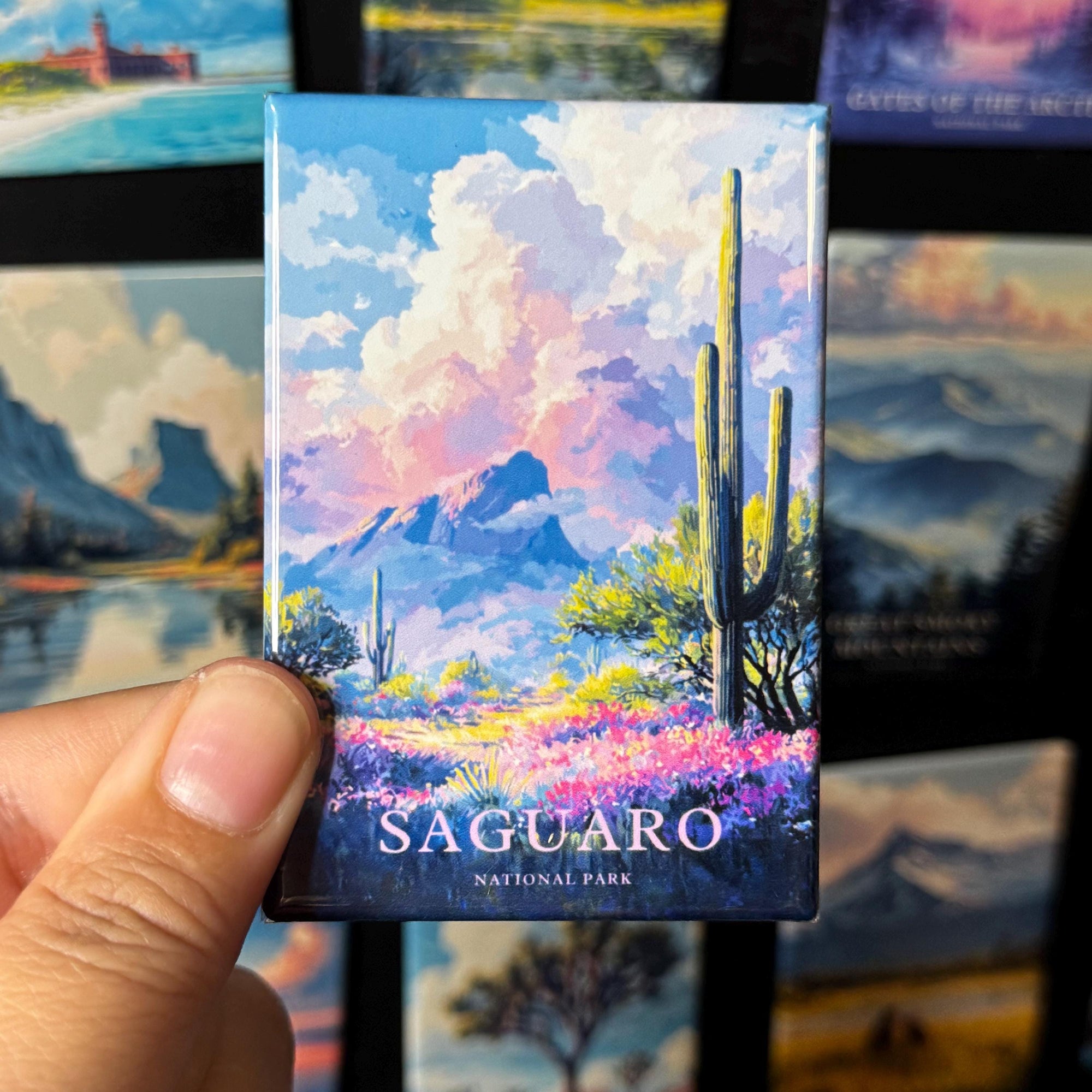 A hand holding a magnet with a colorful illustration of a desert landscape, including mountains, cacti, and flowers.