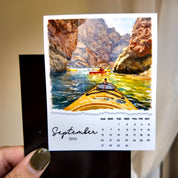 A hand holding a calendar page with a painting of two people kayaking in a canyon.