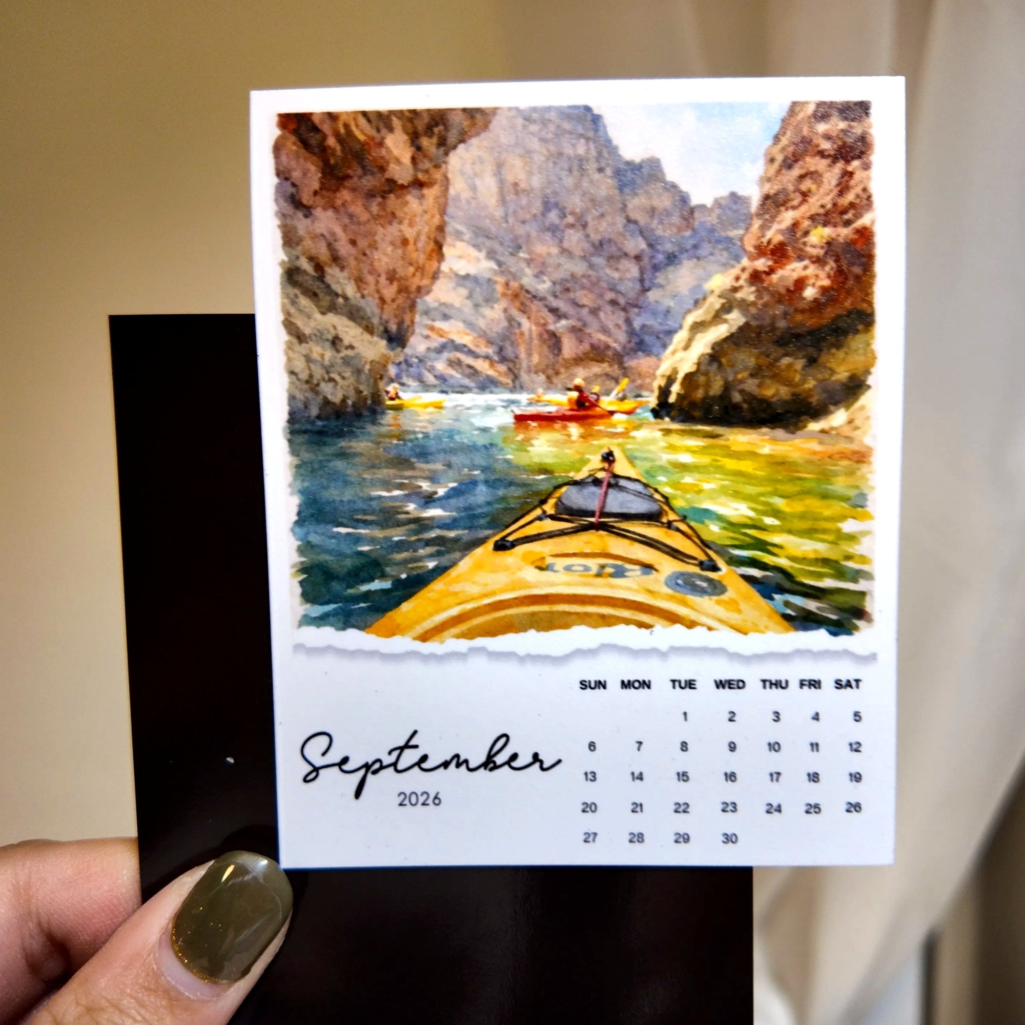 A hand holding a calendar page with a painting of two people kayaking in a canyon.