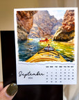 A hand holding a calendar page with a painting of two people kayaking in a canyon.