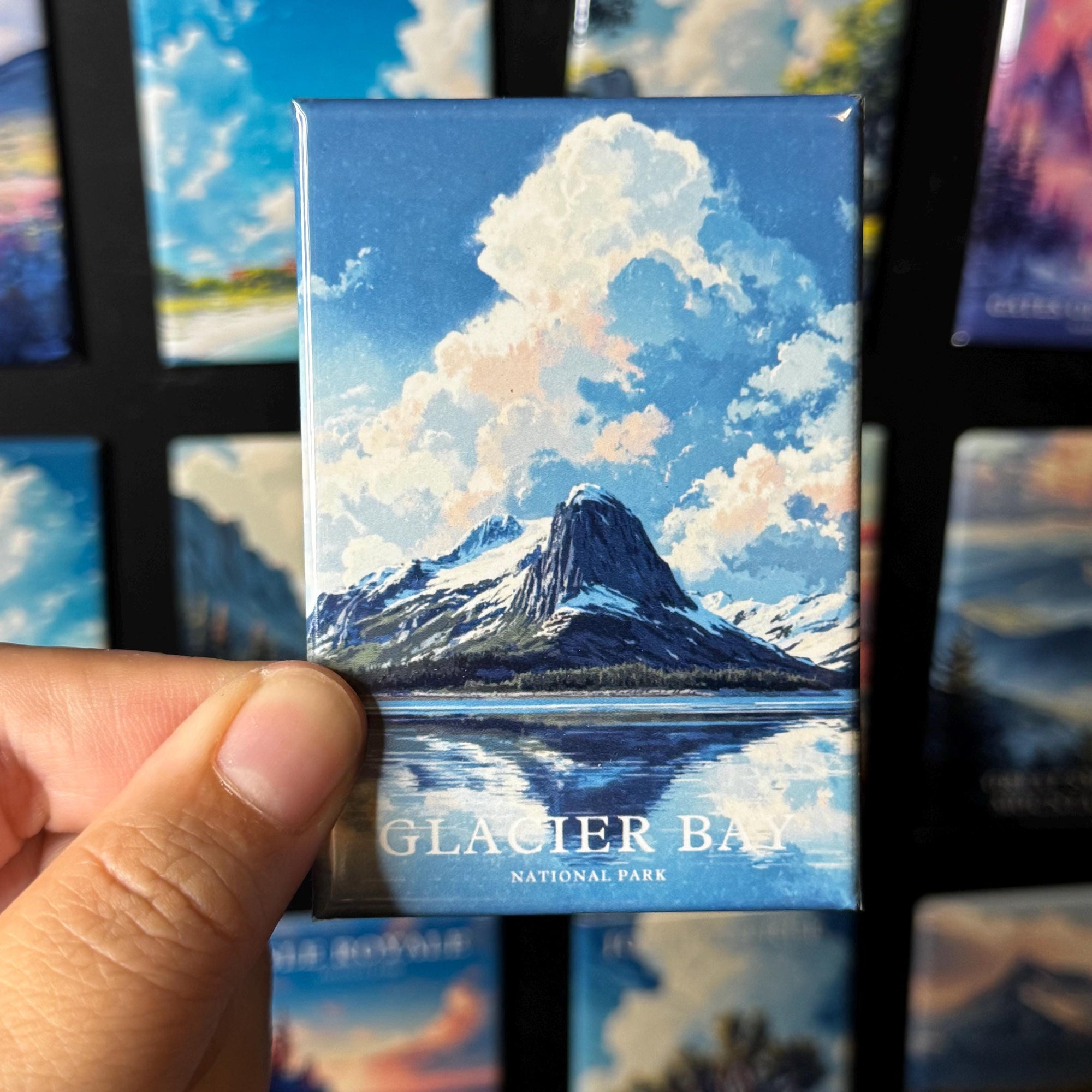 A hand holding a magnet with a picture of a mountain and a lake, with the text "GLACIER BAY NATIONAL PARK" printed on it.