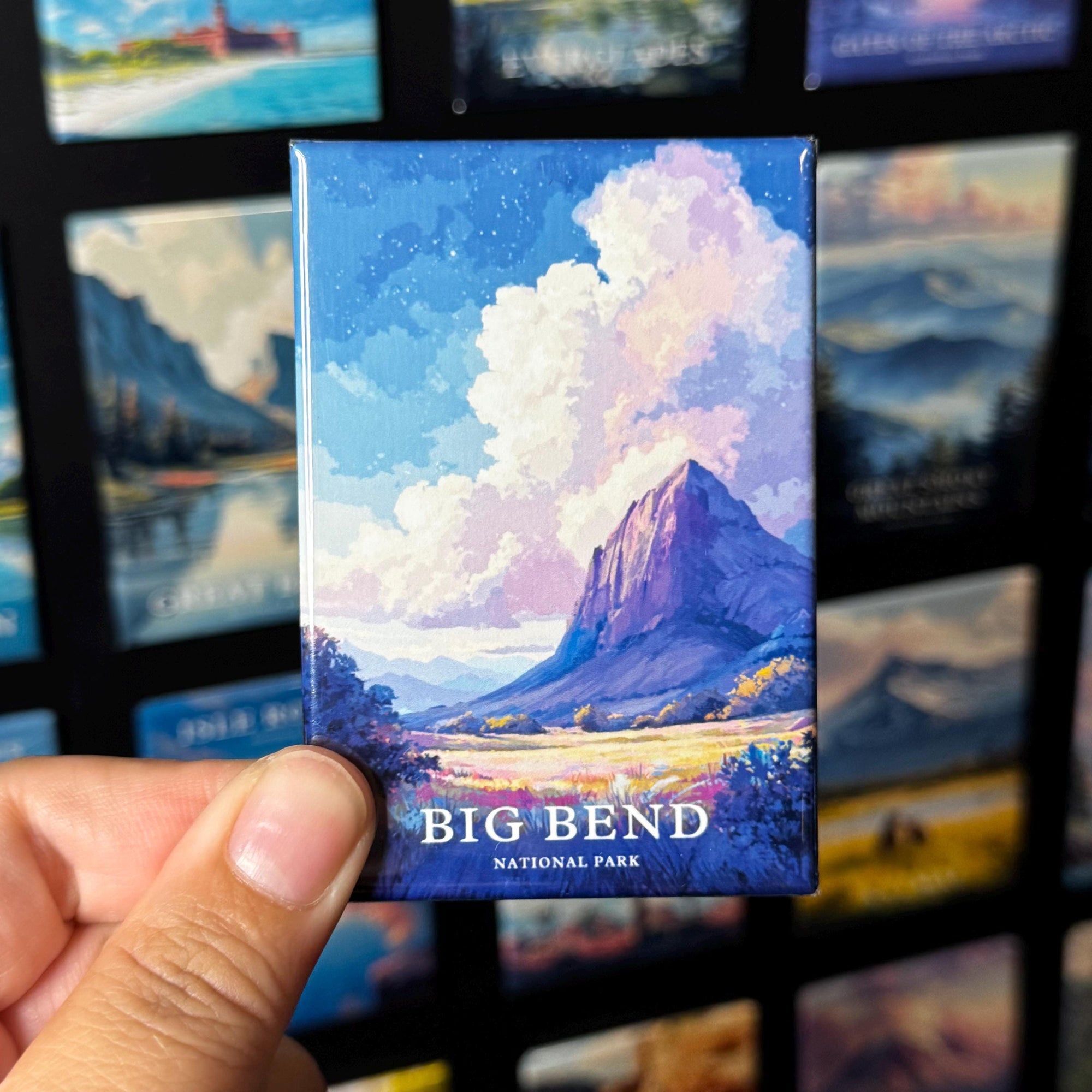 A hand holding a magnet with a landscape scene depicting a mountain and clouds, with the text "BIG BEND NATIONAL PARK" printed on it.