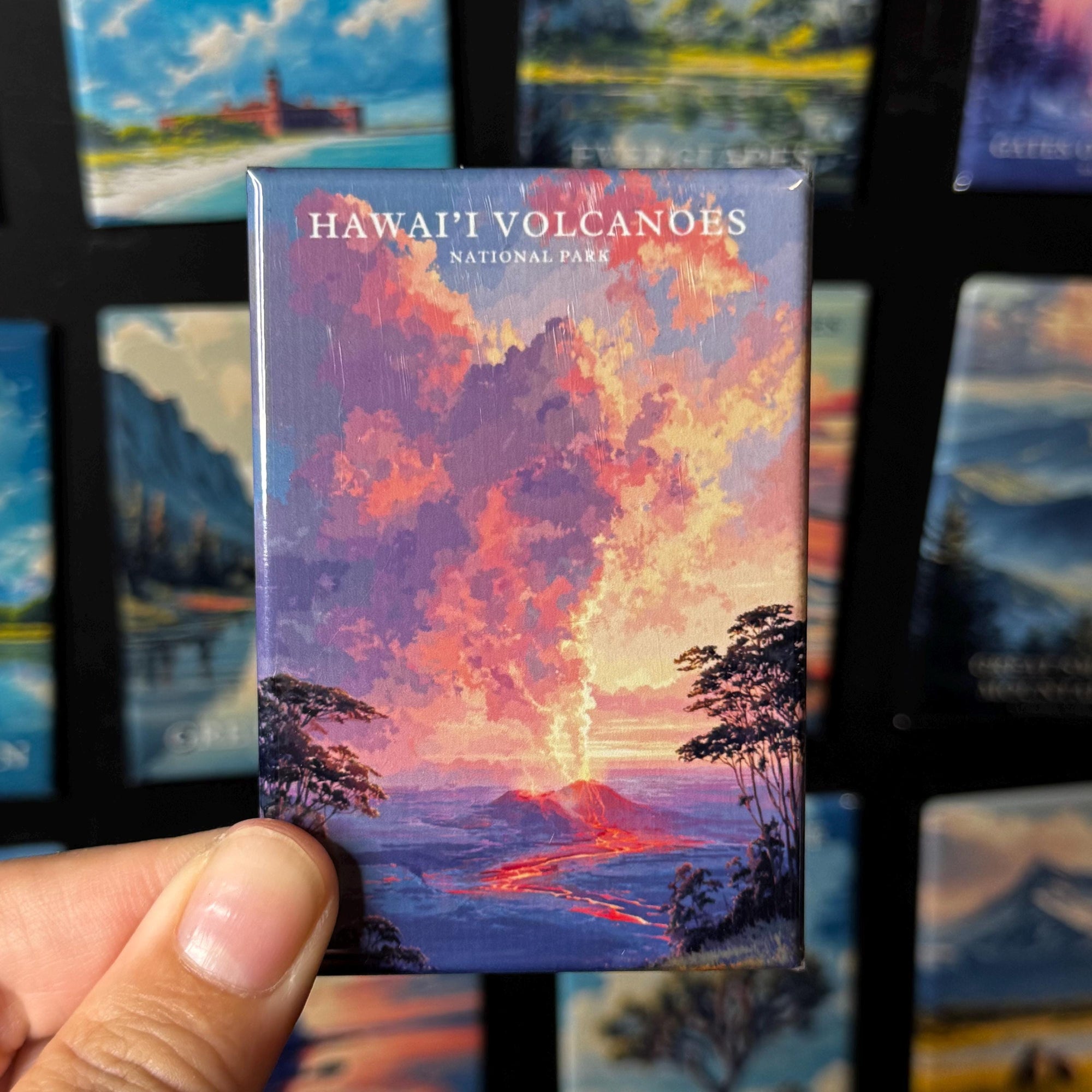 A hand holding a magnet with a colorful illustration of a Hawaiian volcano, with the text "HAWAII' VOLCANOES NATIONAL PARK" printed on it.