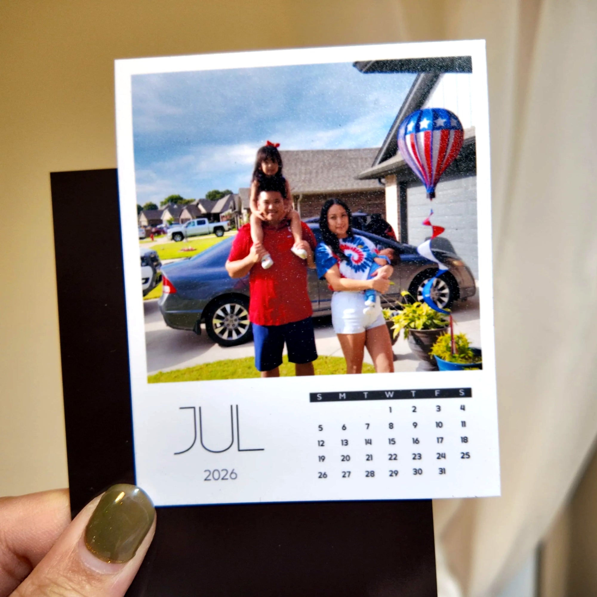 A person is holding a small calendar with a photo of a family standing in front of a house, with a hot air balloon in the background.