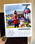 A person is holding a small calendar with a photo of a family standing in front of a house, with a hot air balloon in the background.