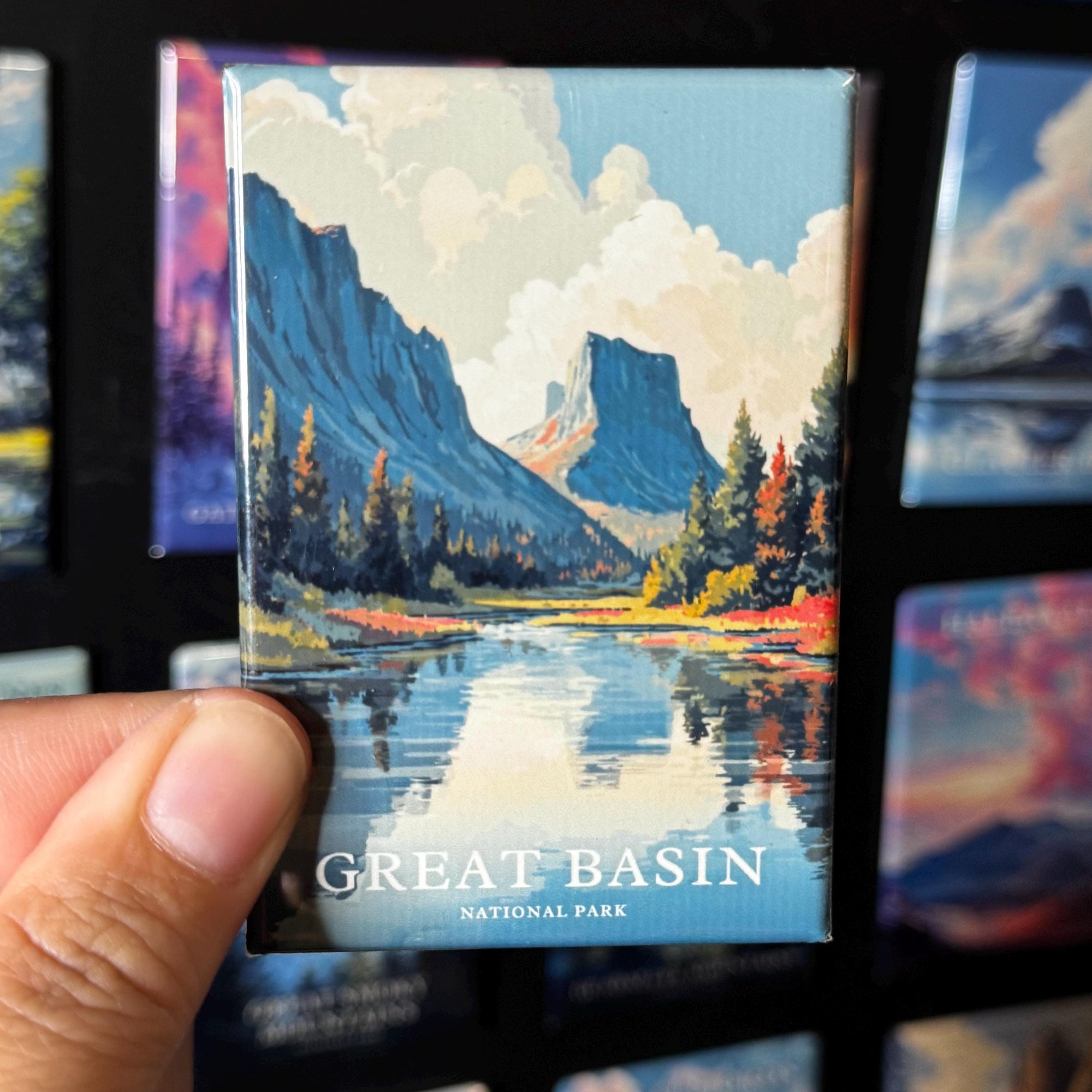 A hand holding a magnet with a painting of the Great Basin National Park, featuring mountains, trees, and a river.
