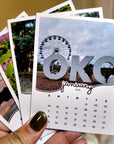 A person's hand holding a calendar with a large "OKC" logo in the center, surrounded by various images of different landscapes and scenes.