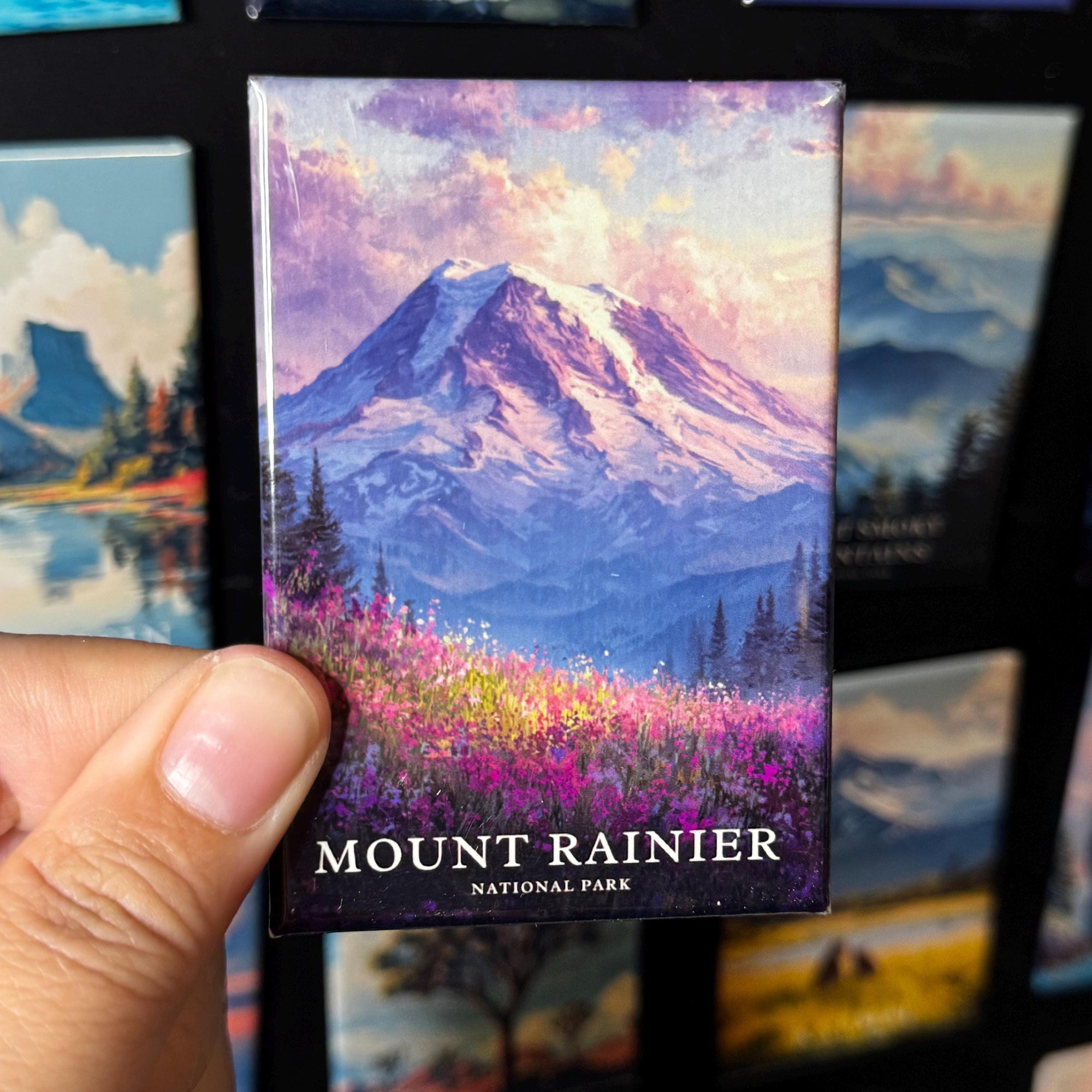 A hand holding a magnet with a painting of a mountain landscape, specifically Mount Rainier, in the background.
