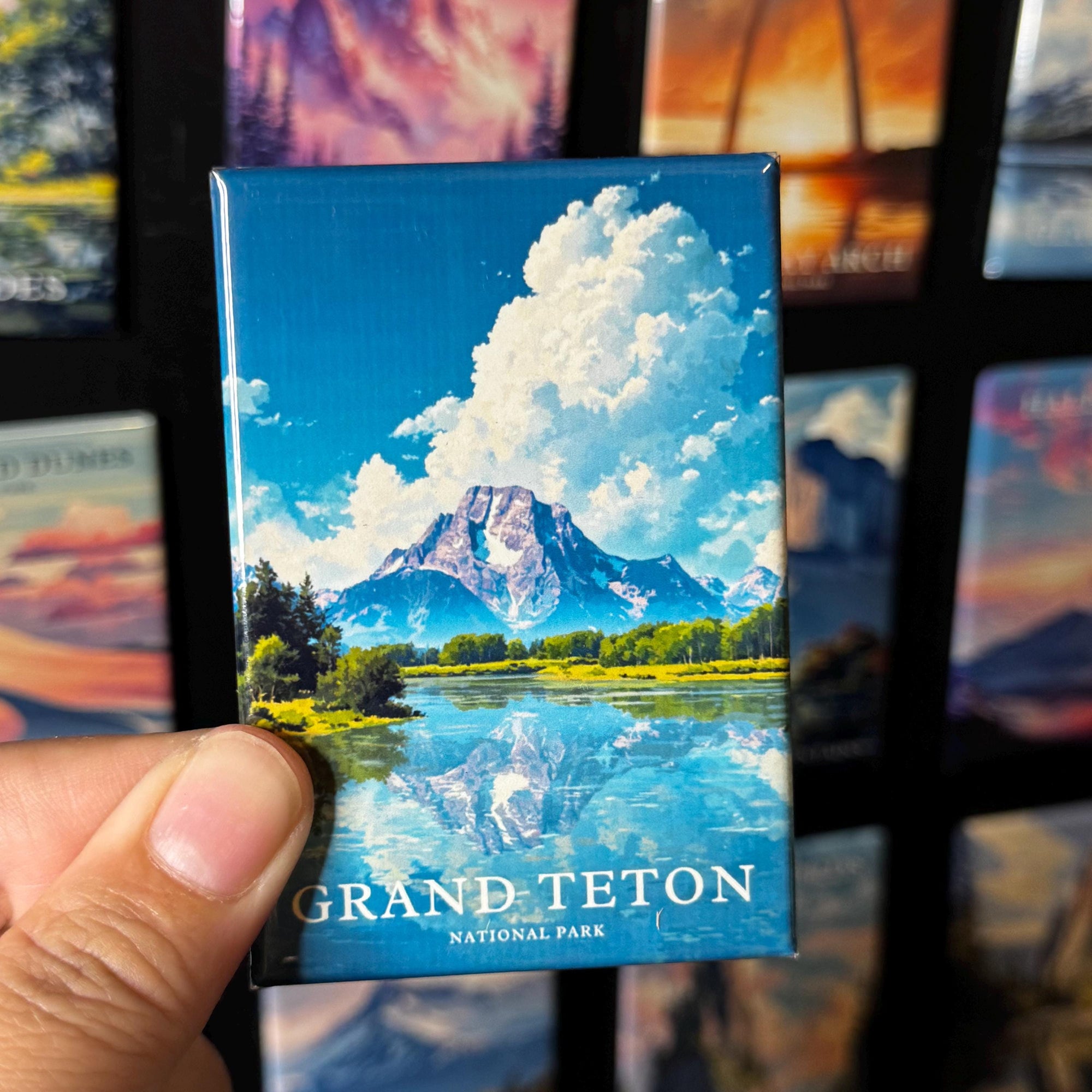 A hand holding a magnet with a scenic image of a mountain and a lake, with the text "GRAND Teton NATIONAL PARK" printed on it.