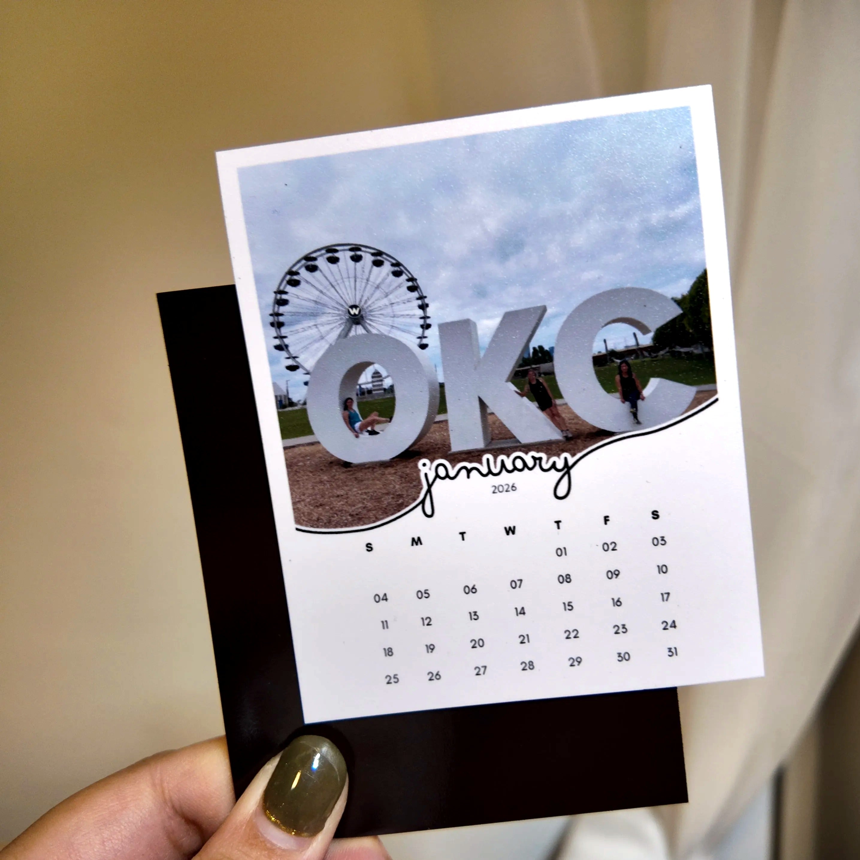 A person is holding a calendar card featuring a large "OKC" logo and a photo of a ferris wheel.
