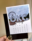 A person is holding a calendar card featuring a large "OKC" logo and a photo of a ferris wheel.