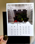 A person is holding a calendar card with a photo of a fireplace and a plant on it.