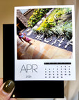 A person holding a small calendar page with a photo of a garden and a calendar for the month of April.