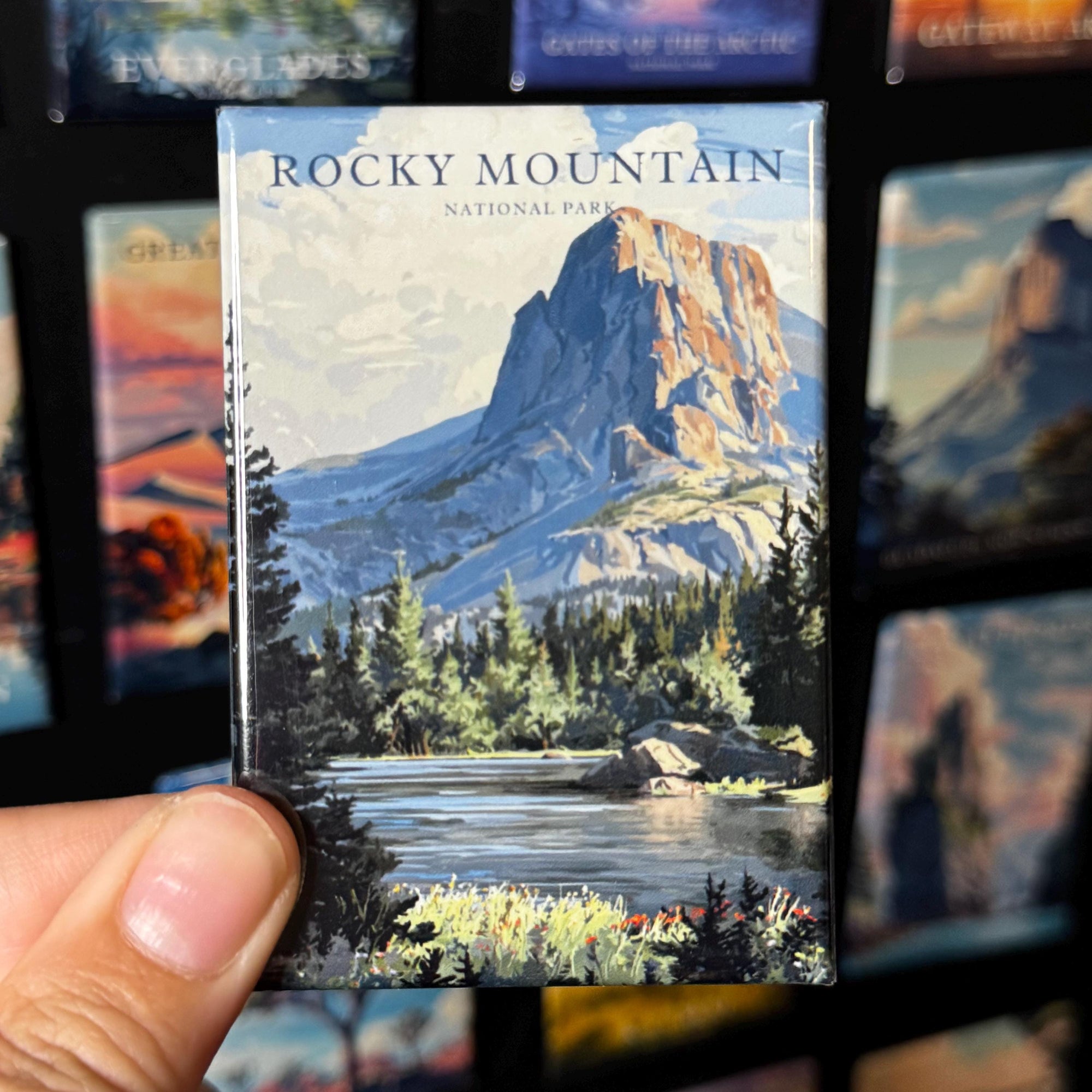 A hand holding a magnet with a picture of Rocky Mountain National Park, featuring a mountain landscape with a lake and forest in the foreground.