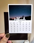 A hand holding a calendar card with a night sky image on it, displaying the month of March and the year 2026.