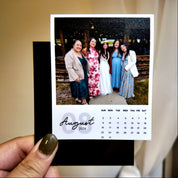 A person is holding a small photo frame containing a printed calendar for the month of August, with a group photo of five women in the background.