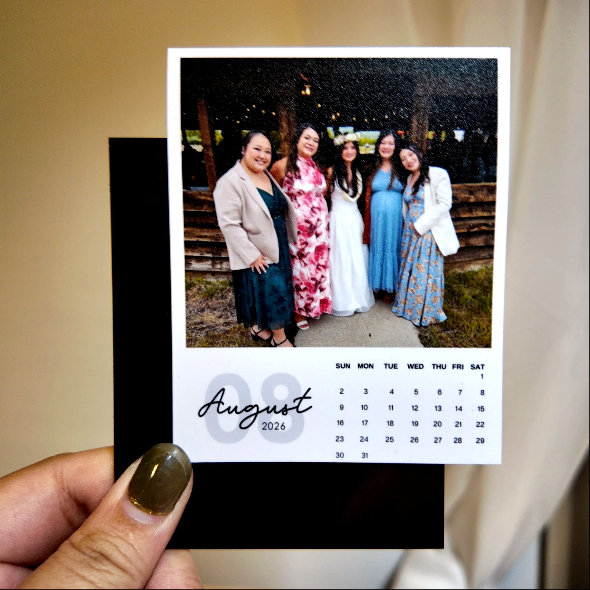 A person is holding a small photo frame containing a printed calendar for the month of August, with a group photo of five women in the background.