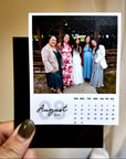 A person is holding a small photo frame containing a printed calendar for the month of August, with a group photo of five women in the background.