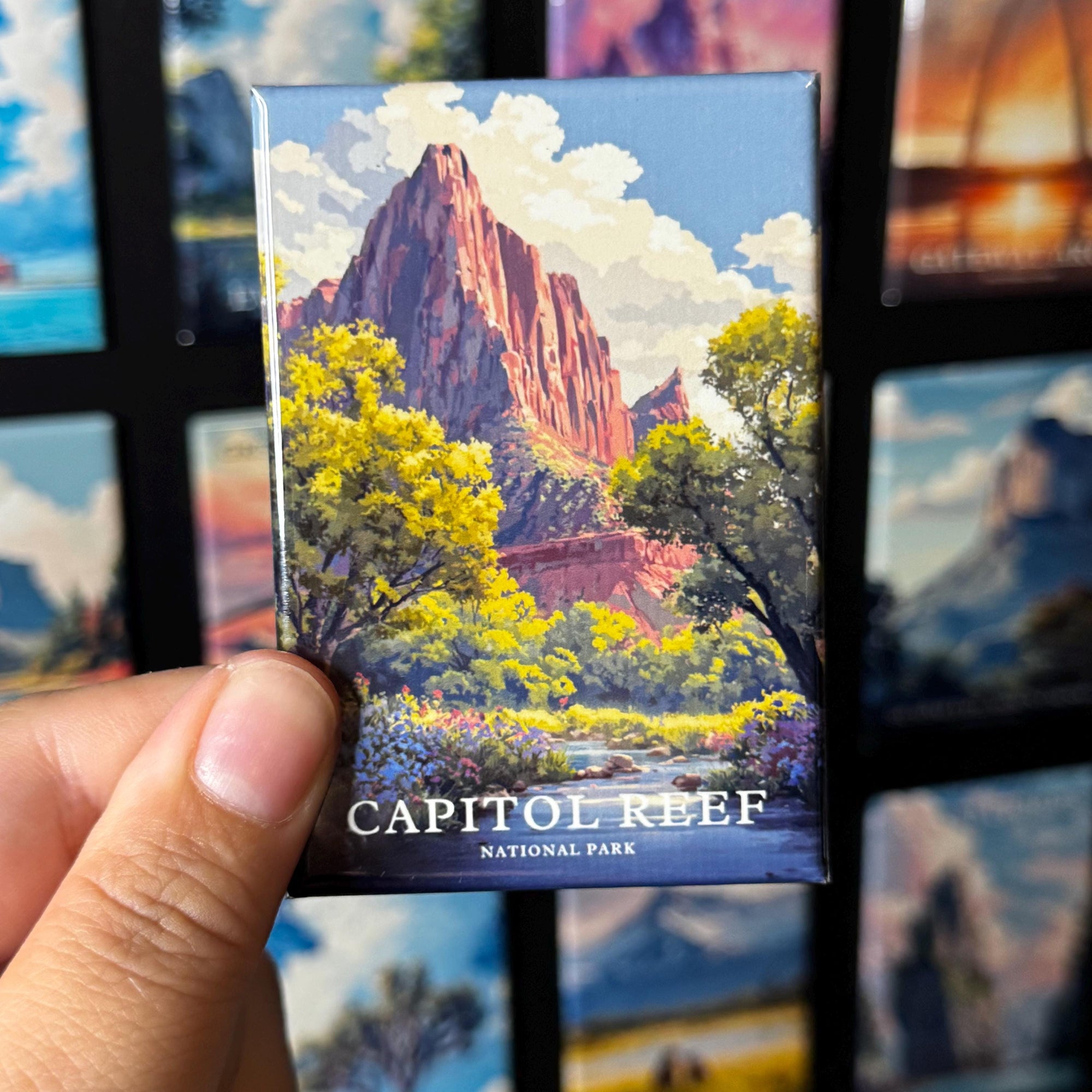A hand holding a magnet with a painting of a mountain landscape, specifically Capitol Reef National Park, against a background of other landscape paintings.