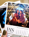 A person's hand holding a calendar with a photo of two women sitting together, with a neon sign visible in the background.