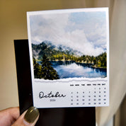 A hand holding a calendar for October 2014, featuring a scenic landscape with a lake and mountains.