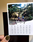 A person is holding a calendar featuring a photo of two people walking on a path through a forest.
